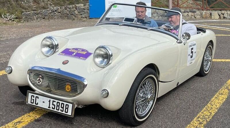 austin healey sprite blanco, parado en curva con fondo montañoso. A bordo, piloto y navegante